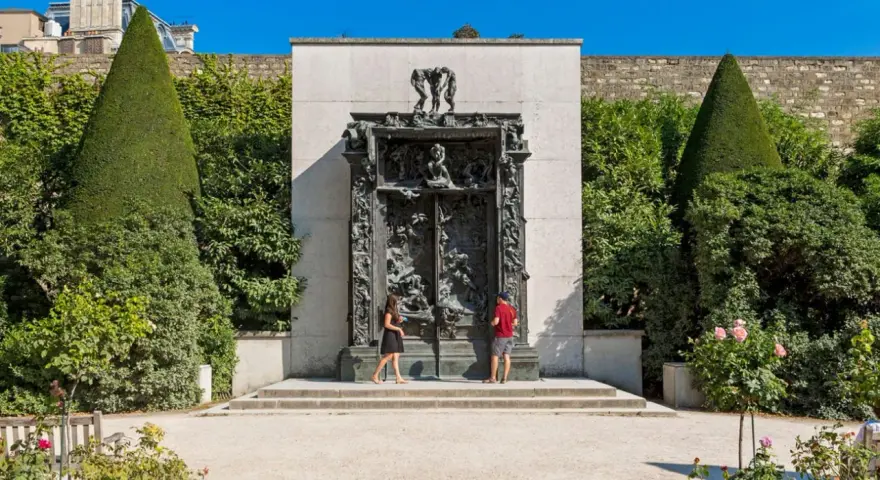 La Porte de l'Enfer, une œuvre monumentale de Rodin, se dresse majestueusement, encadrée par des arbres taillés et un mur de pierre. Deux visiteurs contemplent cette sculpture complexe.