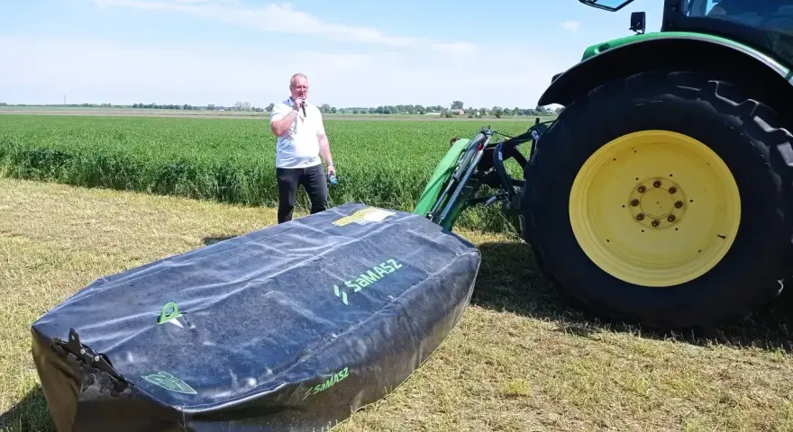 Mężczyzna prezentuje nowoczesne kosiarki dyskowe SaMASZ obok zielonego traktora na polu.