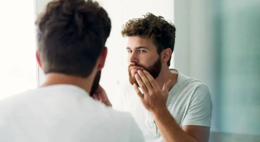 Hombre con barba regia se mira al espejo, acariciando su rostro.