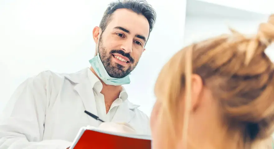 Uśmiechnięty dentysta w białym fartuchu i masce na szyi rozmawia z pacjentką, trzymając notatnik.