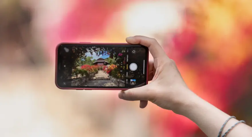 Une main tient un smartphone pour capturer un escalier fleuri menant à un pavillon. L'écran montre les bases de la photographie, avec des options comme "PHOTO" et "PORTRAIT".