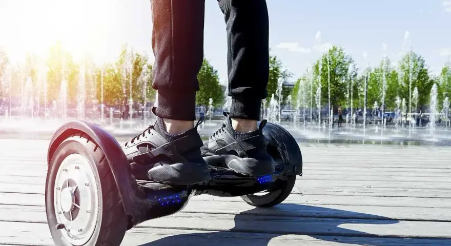 Osoba na deskorolce elektrycznej, czyli urządzeniu transportu osobistego, porusza się po drewnianej promenadzie. W tle widać fontanny i drzewa.