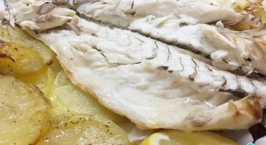 Lubina al horno jugosa y tierna, acompañada de patatas asadas y rodajas de limón fresco.
