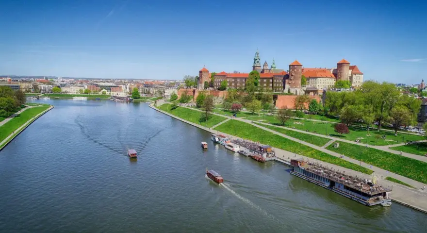 Widok na Wawel i Wisłę. Kilka łodzi pływa po rzece, a na brzegu zielony park i zabudowania.