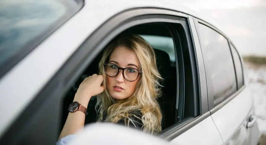 Blondynka w okularach siedzi za kierownicą białego samochodu. Pokazuje, jak nosić okulary, by wyglądać stylowo i pewnie.