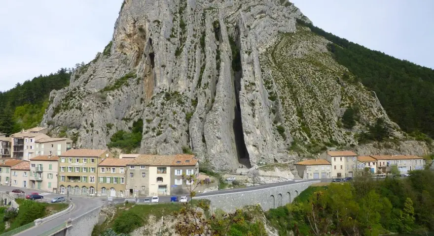 Imponerande klippformation vid Route Napoléon. En by klättrar uppför bergssluttningen, med en bro och en turkos flod nedanför.