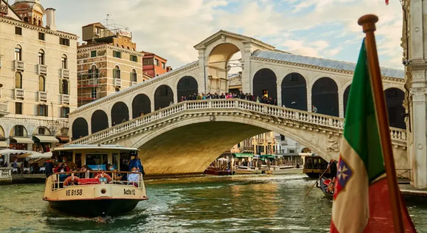 Un vaporetto naviga sul Canal Grande, passando sotto il Ponte di Rialto. L'Italia è rappresentata da una bandiera sventolante.