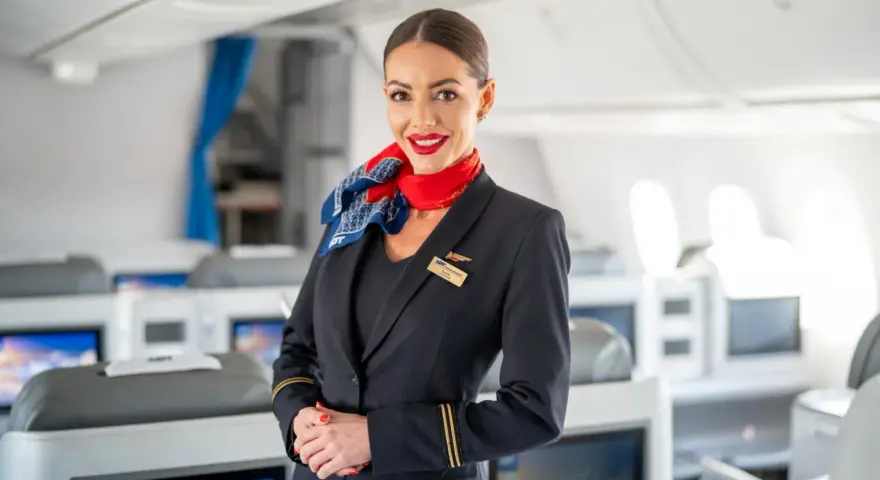 Uśmiechnięta stewardesa w mundurze LOT-u stoi na tle kabiny samolotu z rzędami foteli.