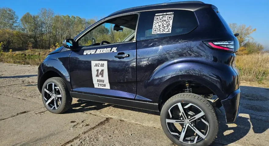 Ciemnoniebieskie Aixam auto z naklejkami "JUŻ OD 14 ROKU ŻYCIA" i kodem QR stoi na tle jesiennego krajobrazu.
