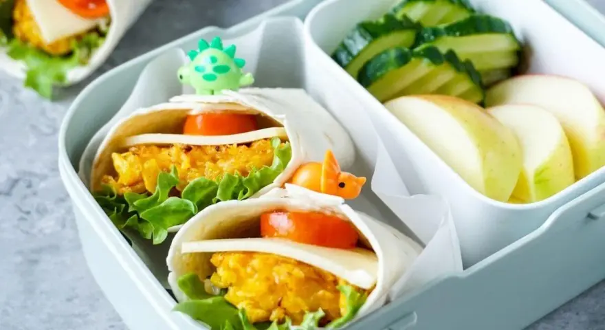 Leckeres Fingerfood für Kinder: Wraps mit Gemüse und Käse, garniert mit kleinen Dino-Spießen. Dazu Gurken und Apfelschnitze.