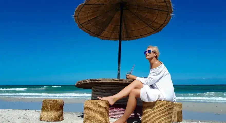 Kobieta w białej sukience relaksuje się pod parasolem na plaży, popijając napój. Idealne tunezja wakacje.
