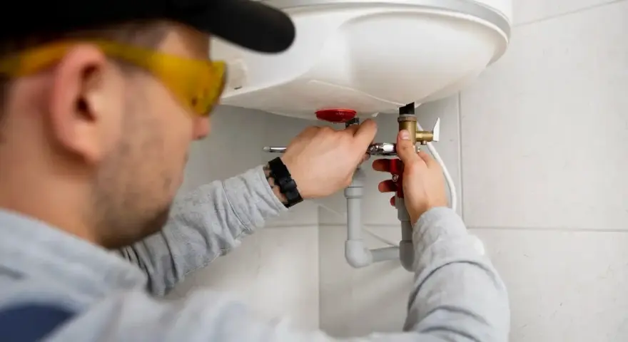 Instalator w okularach ochronnych podłącza bojler elektryczny, pracując przy rurach.