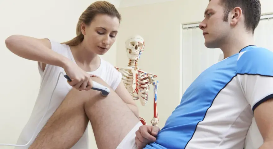 Fizjoterapeutka stosuje urządzenie do terapii, wyjaśniając pacjentowi, co to są bioprądy i jak działają na jego kolano.