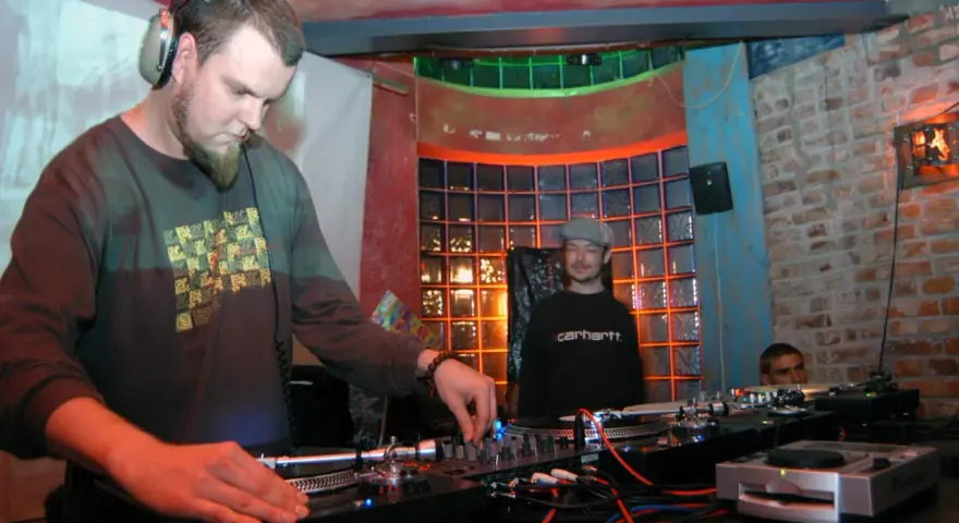 DJ Maceo Wyro w akcji, miksuje muzykę w klubie. W tle widać innych DJ-ów i kolorowe oświetlenie.