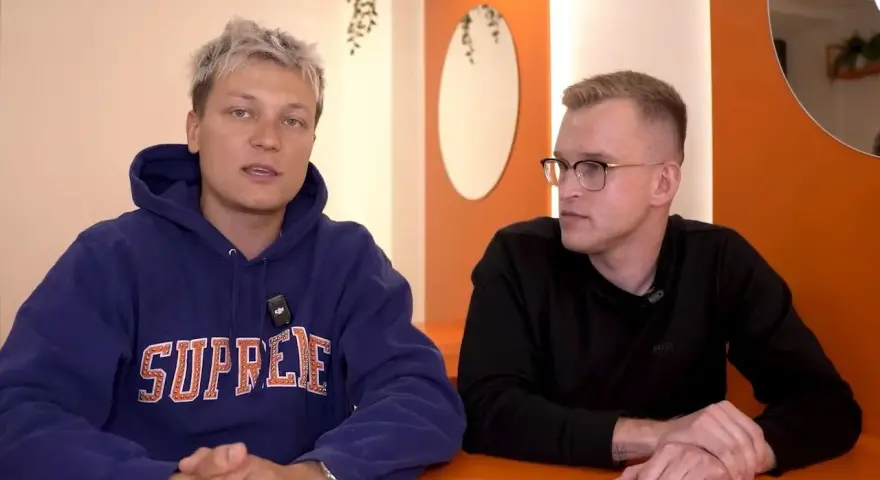 Dwóch mężczyzn siedzi przy pomarańczowym stole. Jeden w niebieskiej bluzie z napisem "SUPREME", drugi w czarnej koszulce. Rozmawiają o kebabie friza.