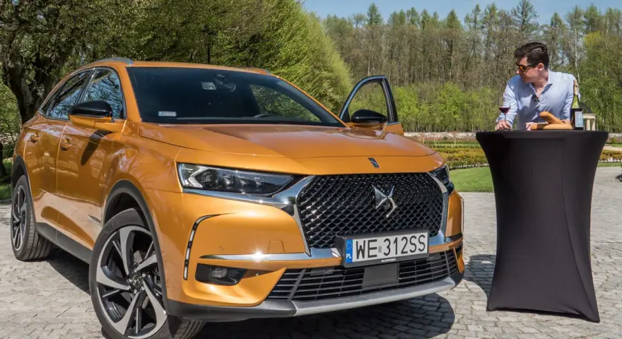 Pomarańczowy DS7 Crossback z otwartymi drzwiami, obok stolik z winem i bagietką. Nawet wady ds7 nie psują uroku tej sceny.