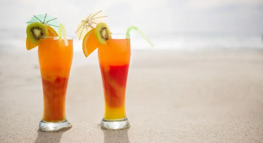Dwa drinki Malibu z sokiem pomarańczowym, ozdobione plasterkami kiwi i pomarańczy, stoją na piaszczystej plaży.