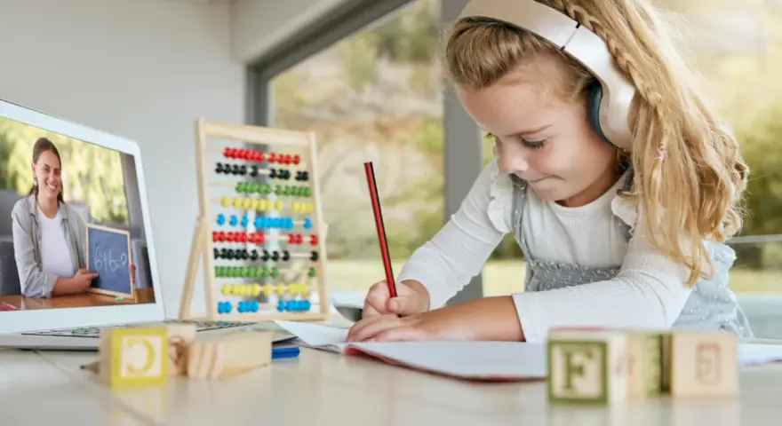 Dziewczynka w słuchawkach uczy się matematyki online, pisząc w zeszycie. Na ekranie laptopa nauczycielka pokazuje zadanie.