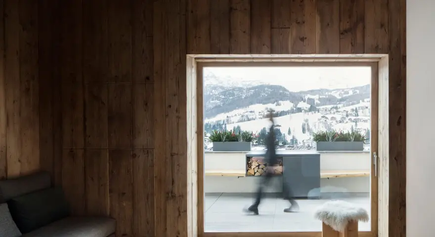 Interni di case di montagna con pareti in legno scuro, travi a vista e un divano grigio. Fuori, un paesaggio innevato.