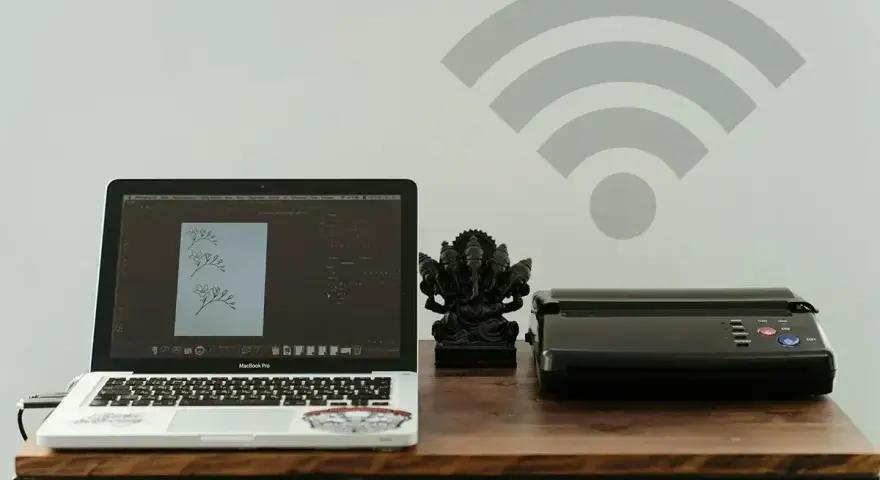 Laptop z projektem graficznym i drukarka obok figurki Ganesha. Symbol Wi-Fi nad nimi sugeruje bezprzewodowe drukowanie.