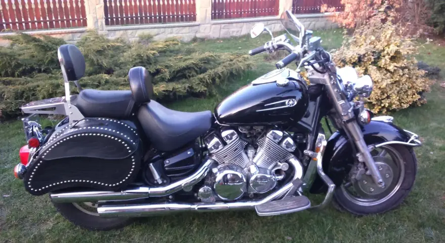 Czarny motocykl Yamaha Royal Star z chromowanymi detalami i skórzanymi sakwami, zaparkowany na trawie przed płotem.