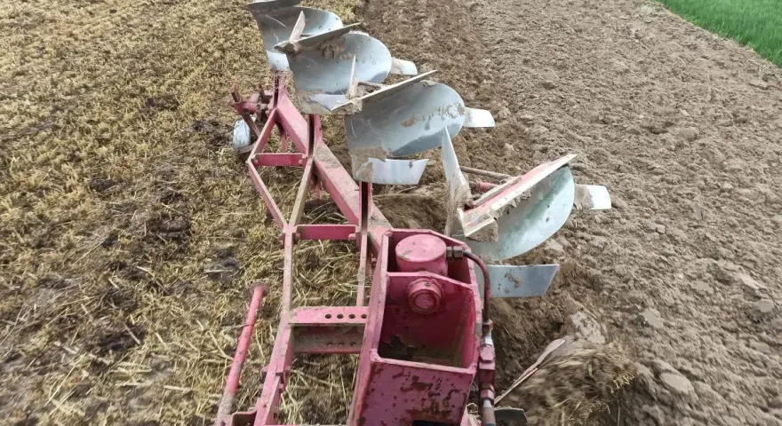 Czerwony pług z kilkoma lemieszami pracuje na polu, odwracając ziemię. Trwa regulacja pługa, przygotowując pole do siewu.