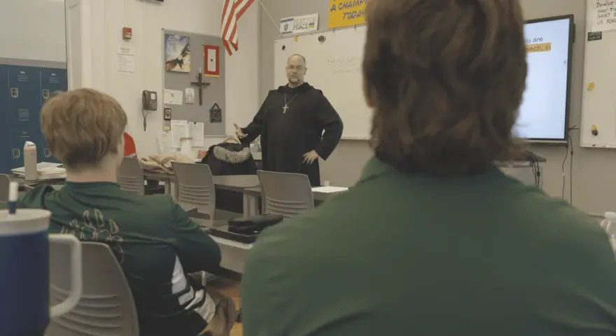 A monastery leader in black robes addresses students in a classroom.