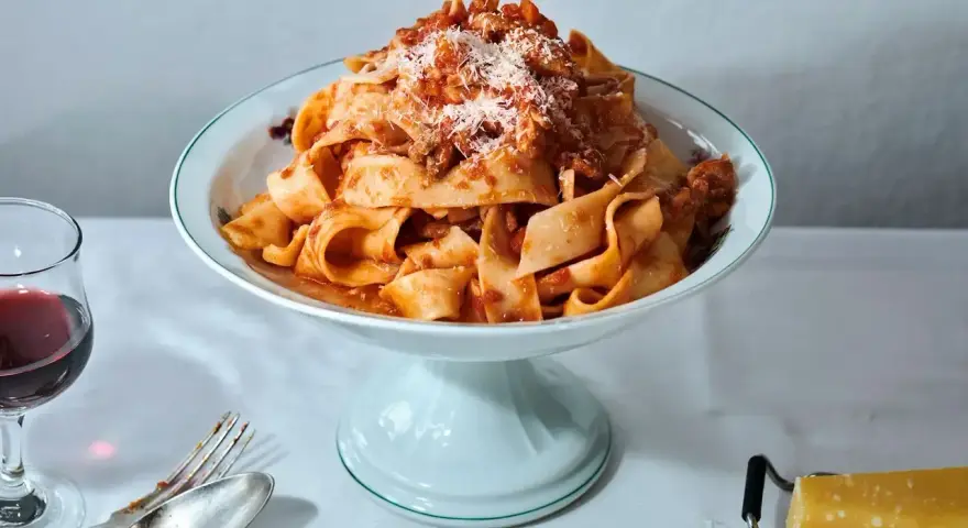 Breite Bandnudeln mit herzhaftem bolognese ragu, bestreut mit Parmesan. Daneben ein Glas Rotwein und Besteck.