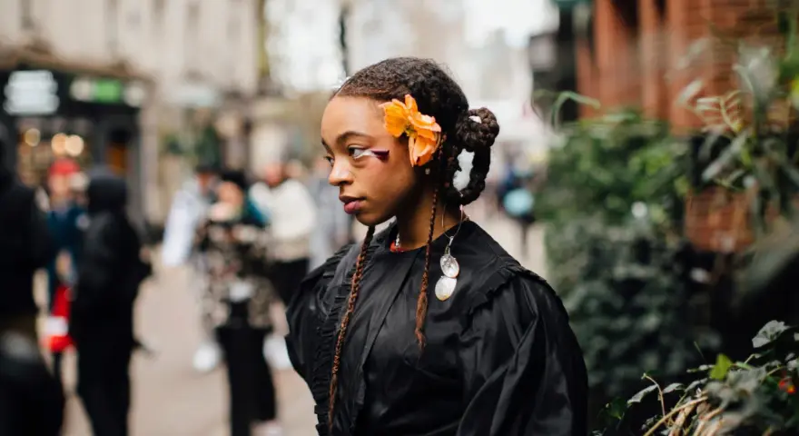 Ragazza con trecce davanti e capelli sciolti, ornata da un fiore arancione e makeup audace. Indossa un abito nero con dettagli voluminosi.
