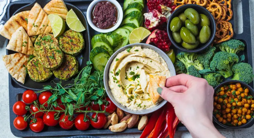 Taca z przekąskami na ognisko: hummus, falafel, warzywa, owoce, pieczywo i paluszki.