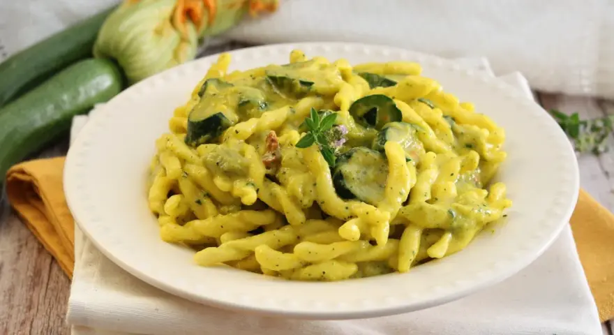 Piatto di pasta con zucchine e zafferano, guarnito con un rametto di erbe aromatiche. Sullo sfondo zucchine fresche e fiori di zucca.