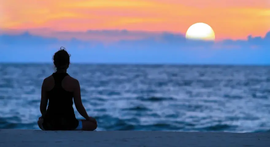En person sitter i lotusställning på en strand och ser ut över havet vid solnedgången. En lugn stund för att meditera hemma, eller var som helst.