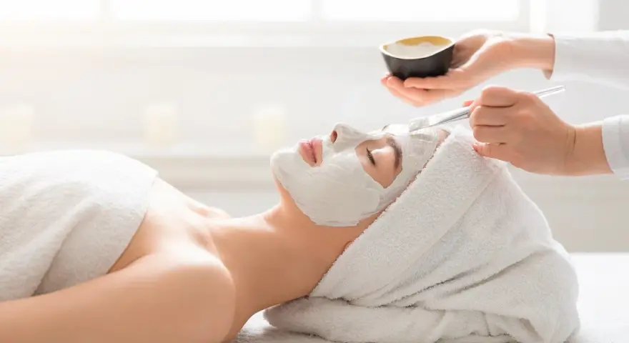 A spa technician applies a white mask to a woman's face. She's wrapped in a towel, enjoying a relaxing facial or massage first.