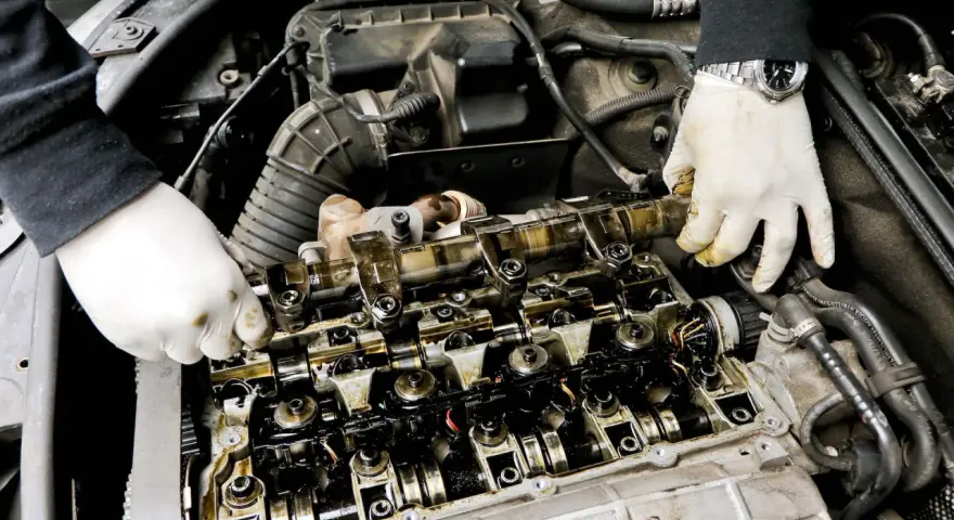 Mechanik w białych rękawiczkach sprawdza wałek rozrządu, szukając objawów zużycia. Widoczne są elementy silnika i przewody.
