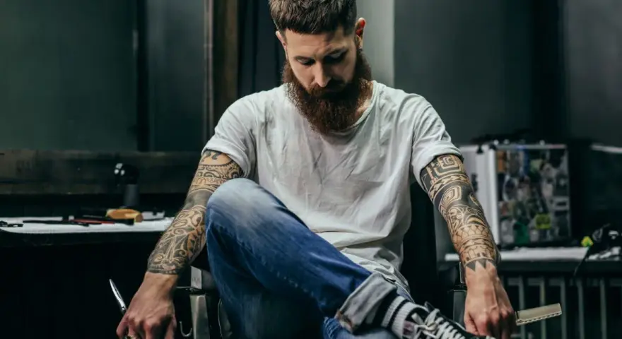 Mężczyzna z brodą i tatuażem na biceps siedzi w fotelu, trzymając nożyczki.