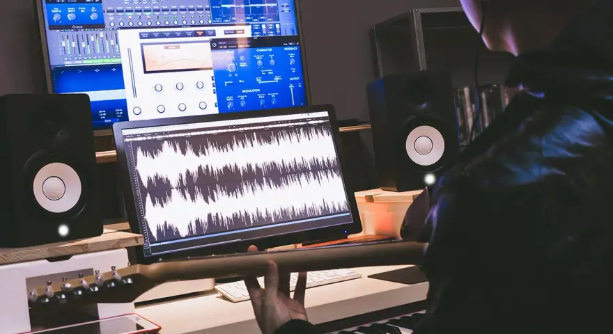 Tworzenie muzyki w domowym studiu. Na ekranach widać programy do edycji dźwięku, a w rękach gitarzysty gryf gitary.
