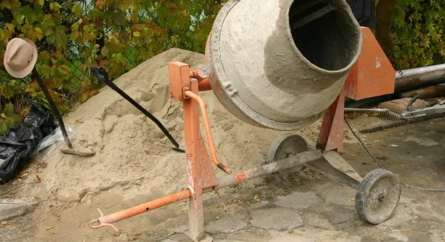 Betoniarka 200l czeka na piasek i cement. Ile cementu na betoniarkę 200l? Obok łopata i kapelusz.