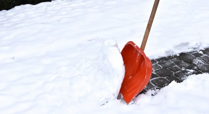 Pomarańczowa łopata do śniegu wbita w zaspę na ośnieżonym chodniku.
