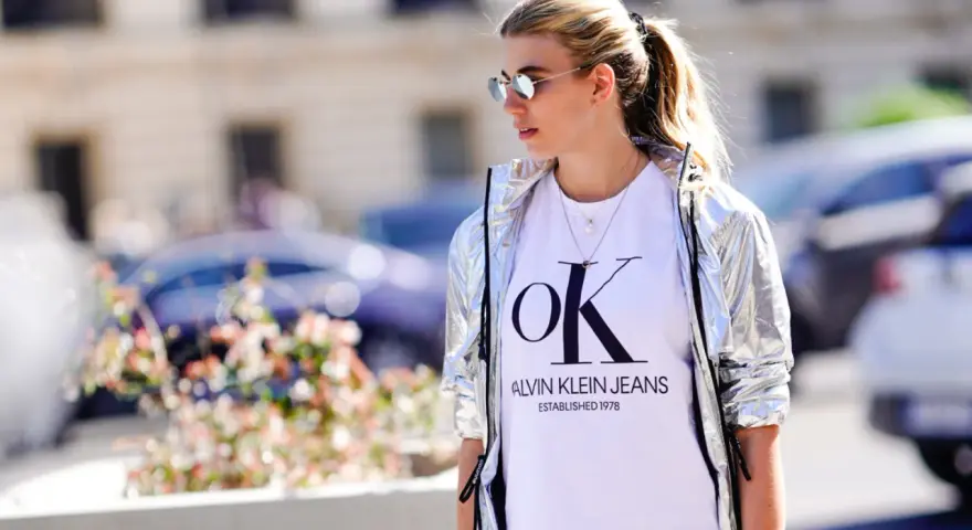 Kobieta w srebrnej kurtce CK damskiej i białej koszulce z logo Calvin Klein Jeans.