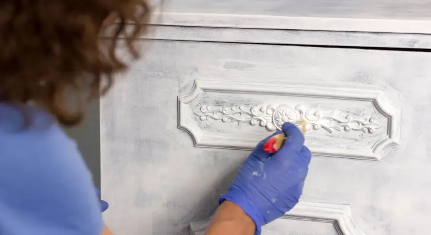 Mano enguantada aplica pintura a muebles pintados a la tiza en dos colores, dando un acabado desgastado y elegante.