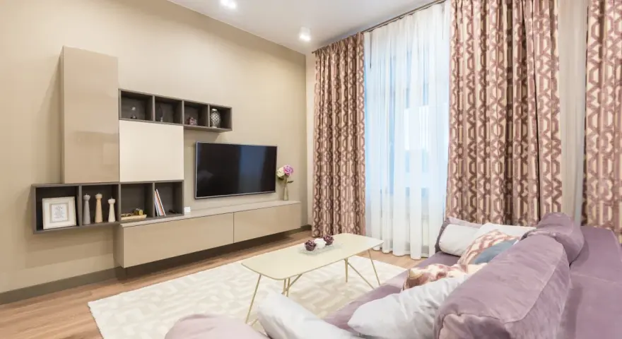 Przytulny salon z fioletową kanapą, beżowy kolor ścian tworzy ciepłą atmosferę. W tle telewizor i zasłony.