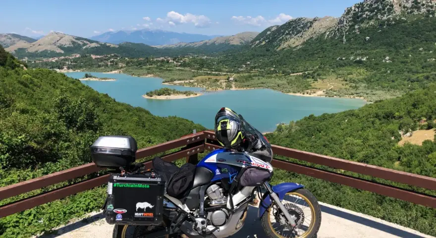 Moto pronta per avventurosi giri in moto nel Lazio, con un panorama mozzafiato sul lago turchese e le montagne verdi.