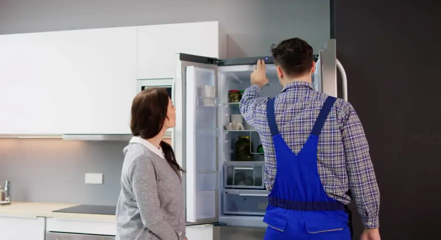 Un technicien en salopette bleue vérifie un frigo qui claque, tandis qu'une femme observe attentivement.