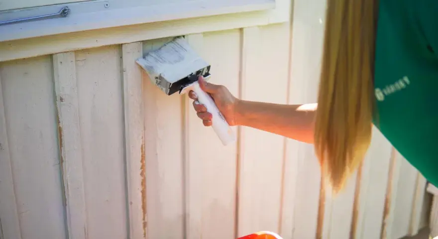 En person med långt blont hår målar en vit träfasad med en bred pensel. Det är dags att måla av huset för sommaren.