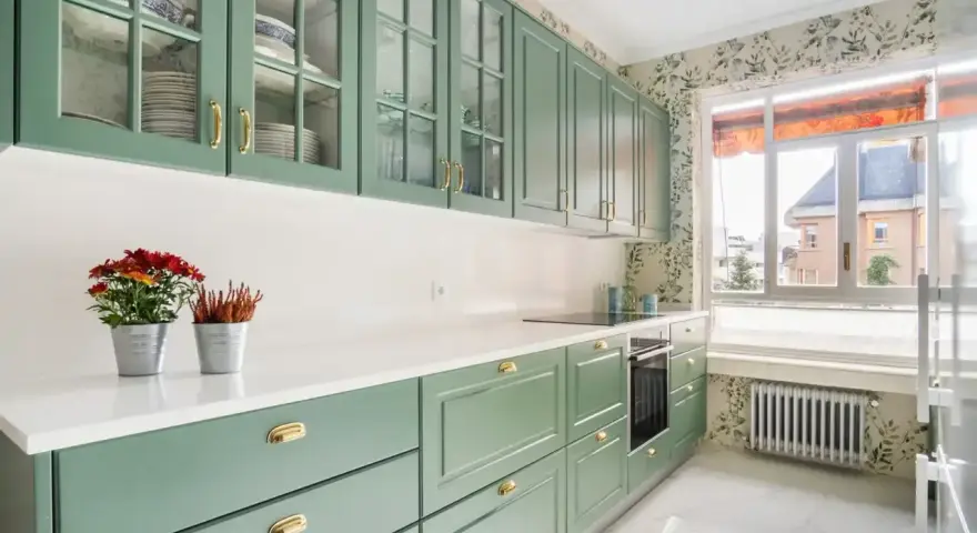 Encantadoras cocinas verdes vintage con armarios de cristal y detalles dorados. Flores rojas y naranjas adornan la encimera blanca.