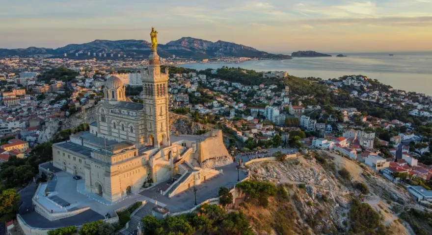 La basilique Notre-Dame de la Garde veille sur Marseille, baignée par la lumière dorée du soleil couchant. La mer et les collines environnantes s'étendent à perte de vue.