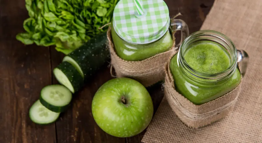 Dwa słoiki z zielonym smoothie, obok jabłko, plasterki ogórka i sałata. Orzeźwiający drink z ogórkiem na drewnianym tle.