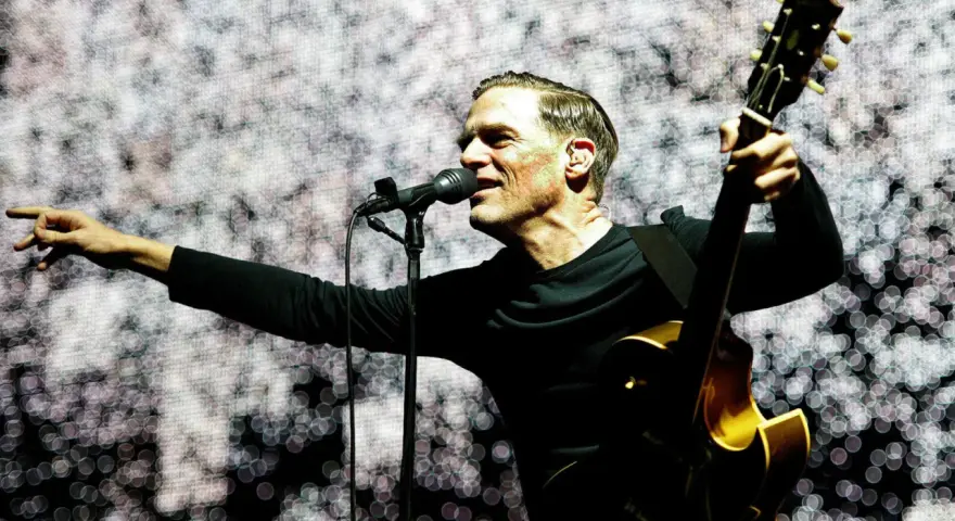 Bryan Adams w Polsce, śpiewa i gra na gitarze na tle świetlistej scenografii.