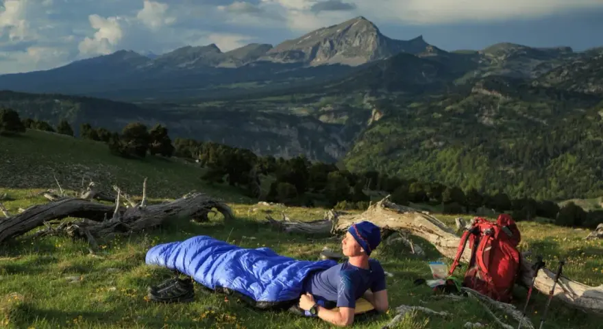 Mann liegt in blauer Biwakschachtel im Freien, umgeben von Berglandschaft. Rucksack und Wanderstöcke daneben.