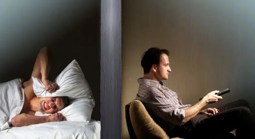 Hombre en cama tapa oídos con almohada, sufriendo por vecinos ruidosos. Al otro lado, otro hombre ve TV.